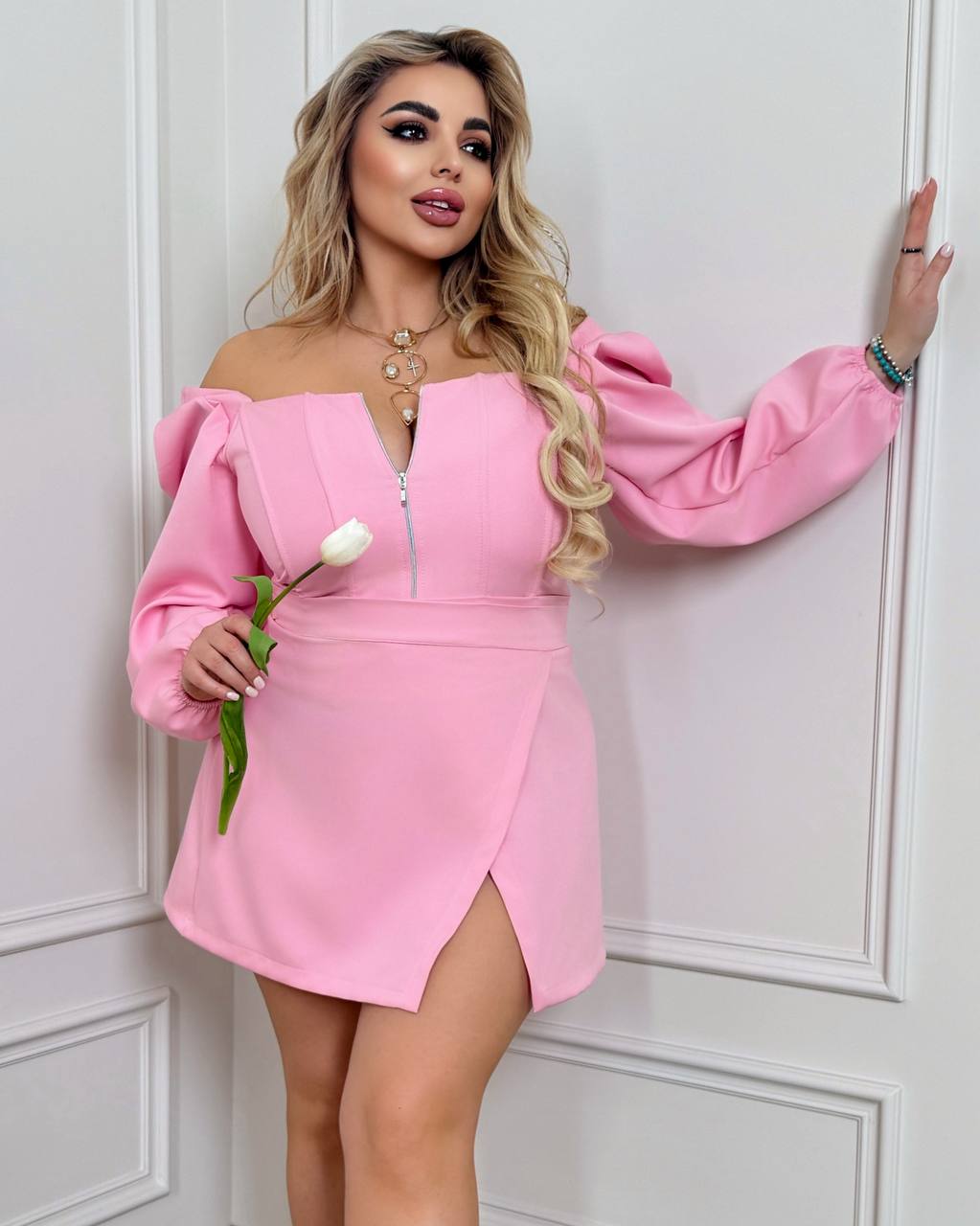 Woman wearing a pink off-shoulder dress holding a white tulip against a white wall.