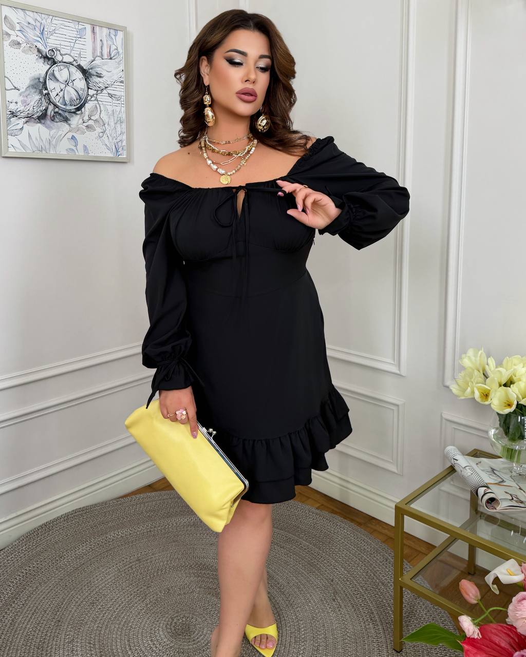 Woman in a black off-shoulder dress holding a yellow clutch in an elegant room.