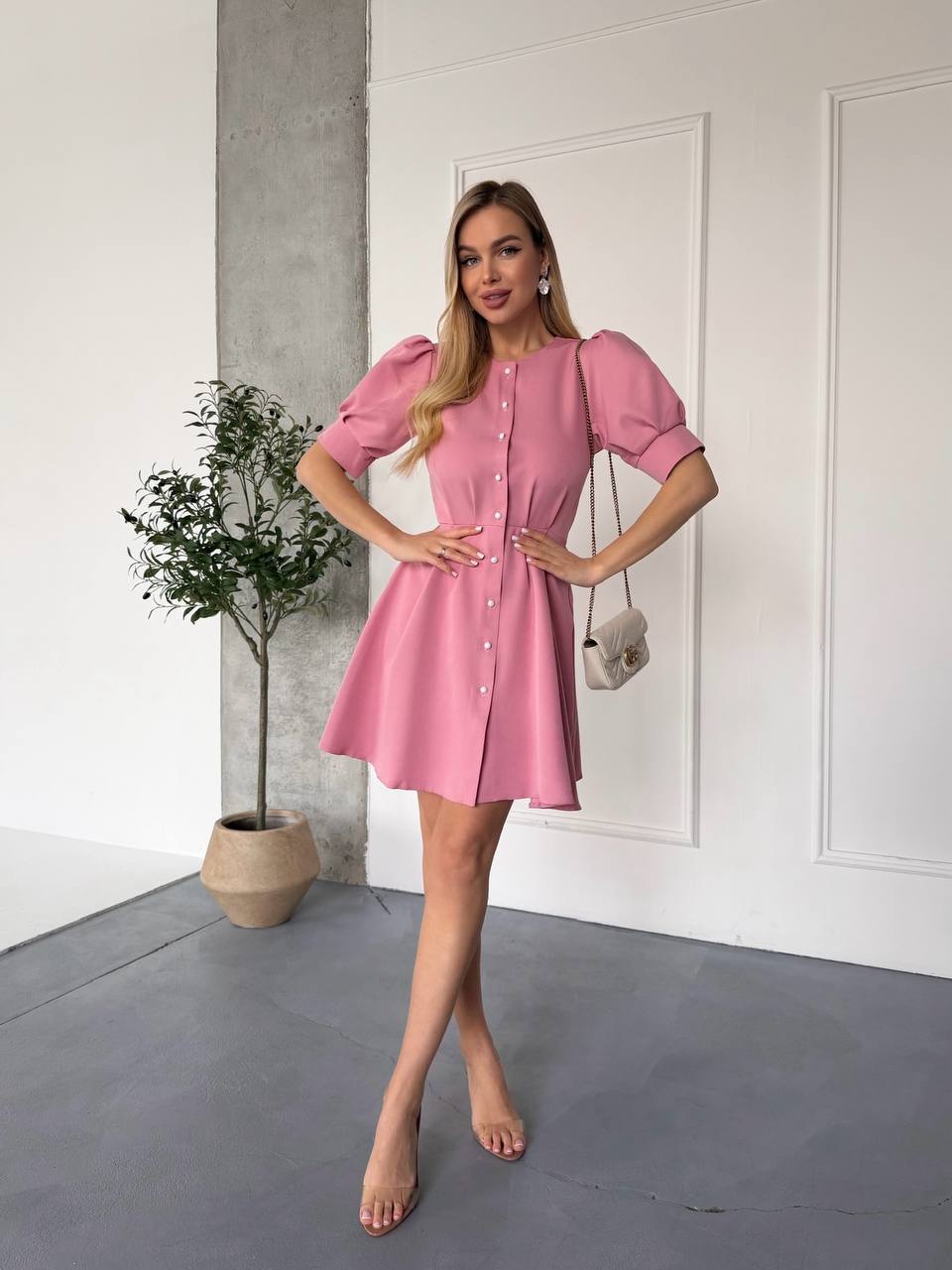 Woman wearing a pink dress standing in a minimalistic room with a plant and white wall.