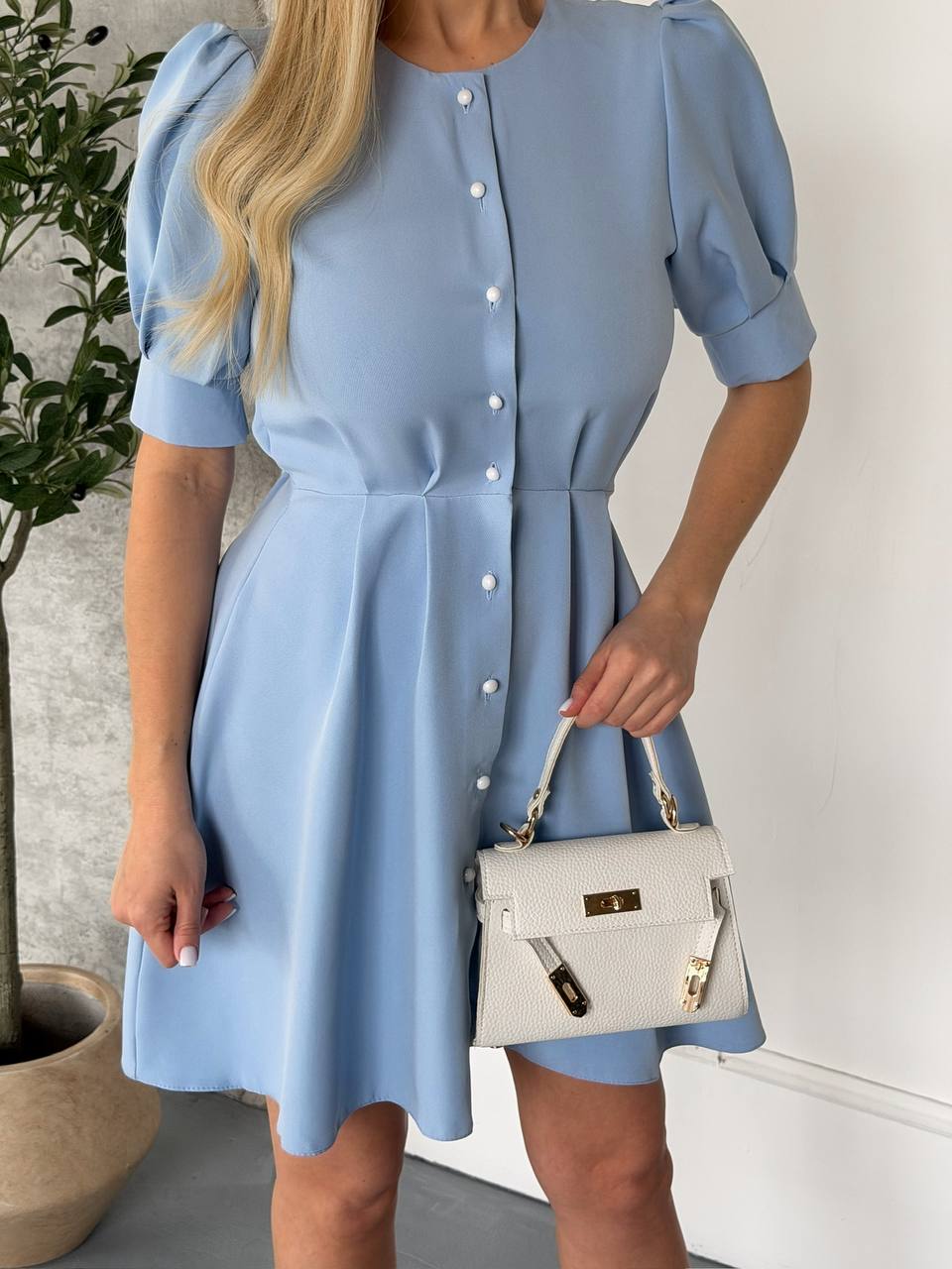 Woman wearing a light blue dress holding a white handbag indoors.