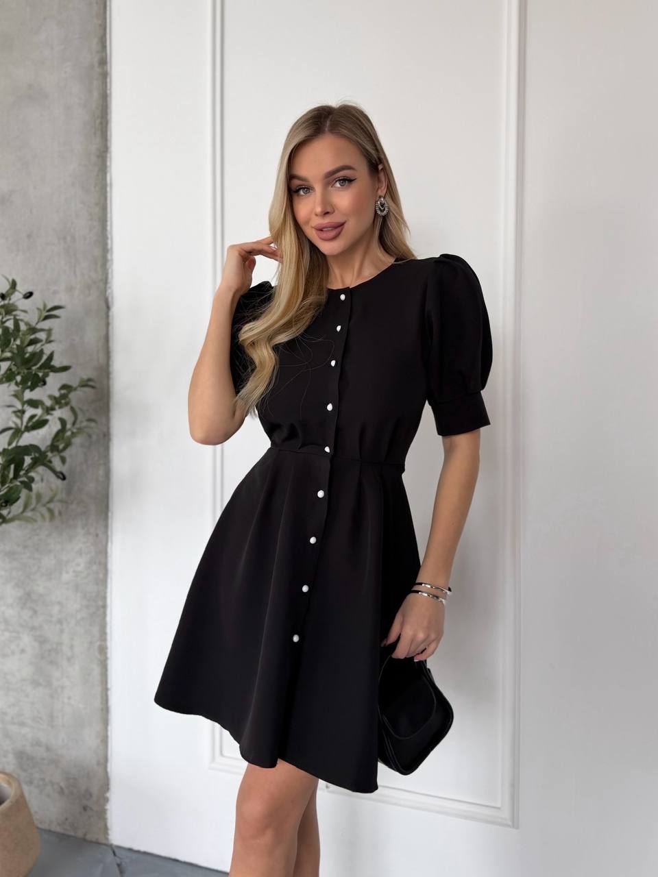 Woman wearing a black dress with white buttons against a white wall.