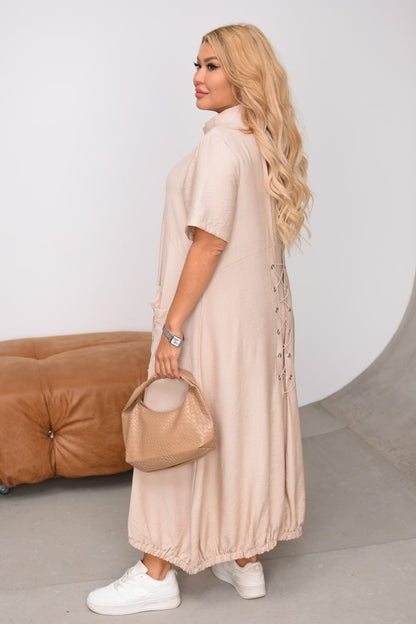 Woman in a long beige dress holding a matching handbag against a neutral background