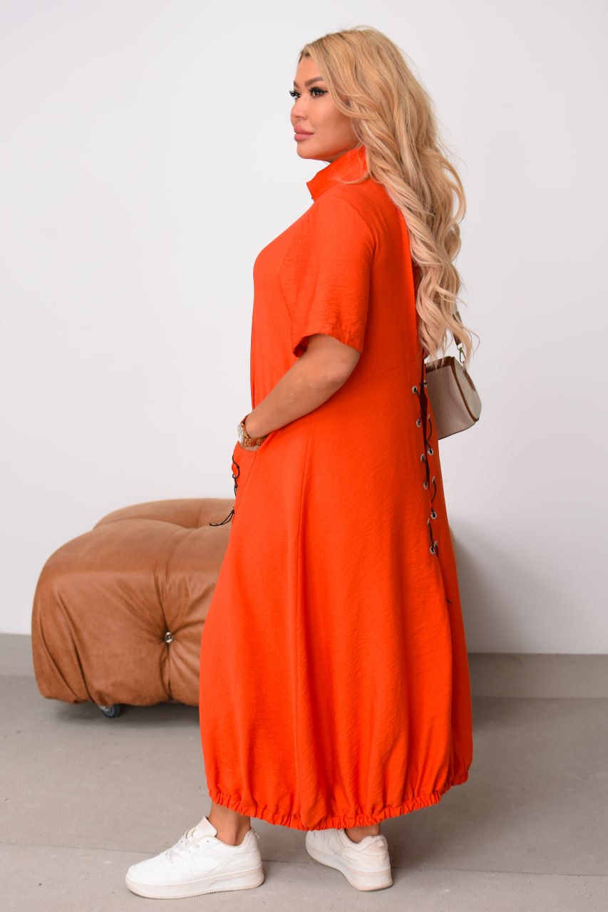 Woman wearing an orange dress standing against a white wall with a brown chair in the background.