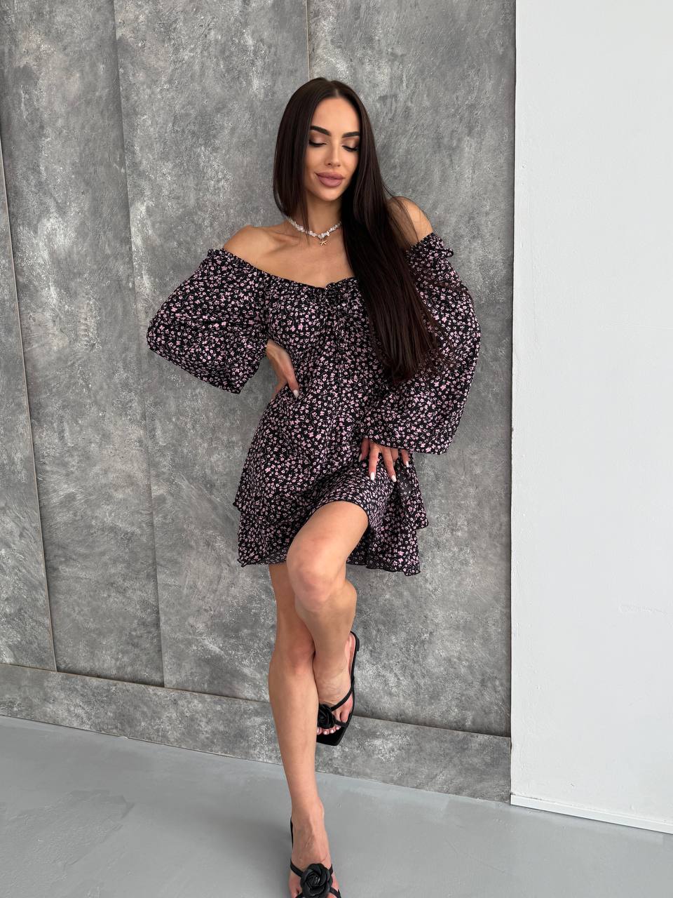 Woman wearing a floral off-shoulder dress against a gray wall.
