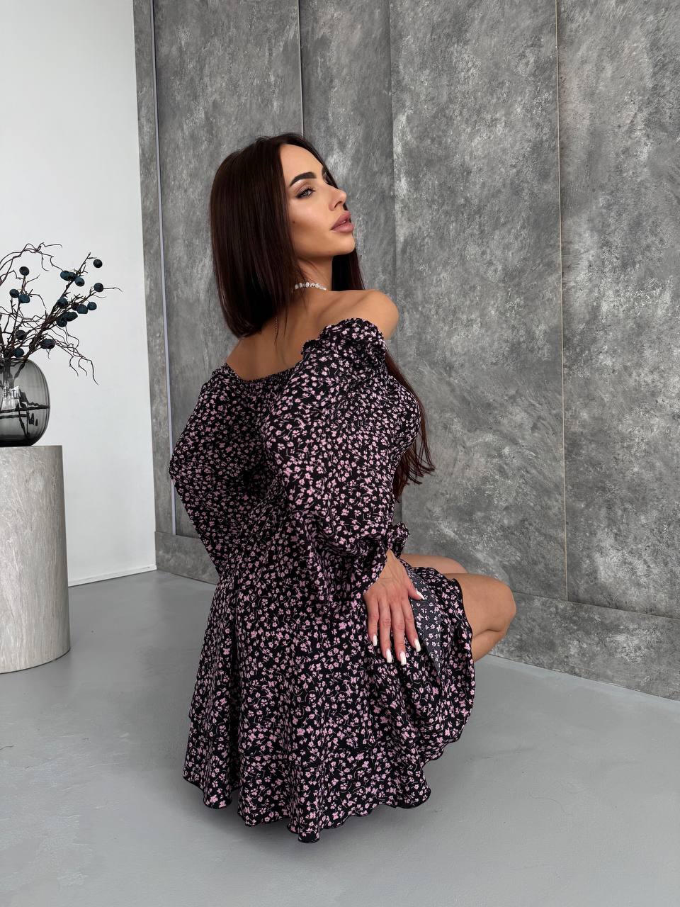 Woman in a floral off-shoulder dress posing against a gray wall.