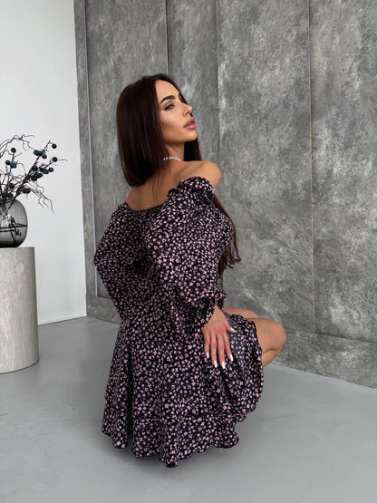 Woman in a floral off-shoulder dress posing against a gray wall.