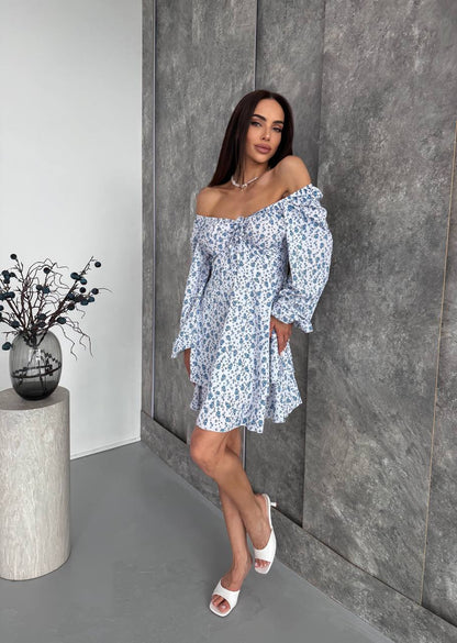 Woman in a floral off-shoulder dress standing against a gray wall with a vase of blackberries on a pedestal.