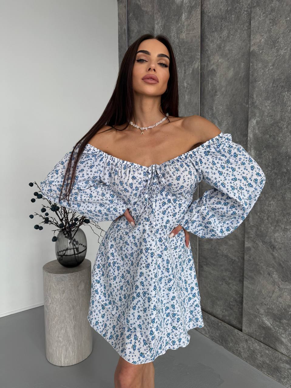 Woman wearing a floral off-shoulder dress standing against a gray wall.
