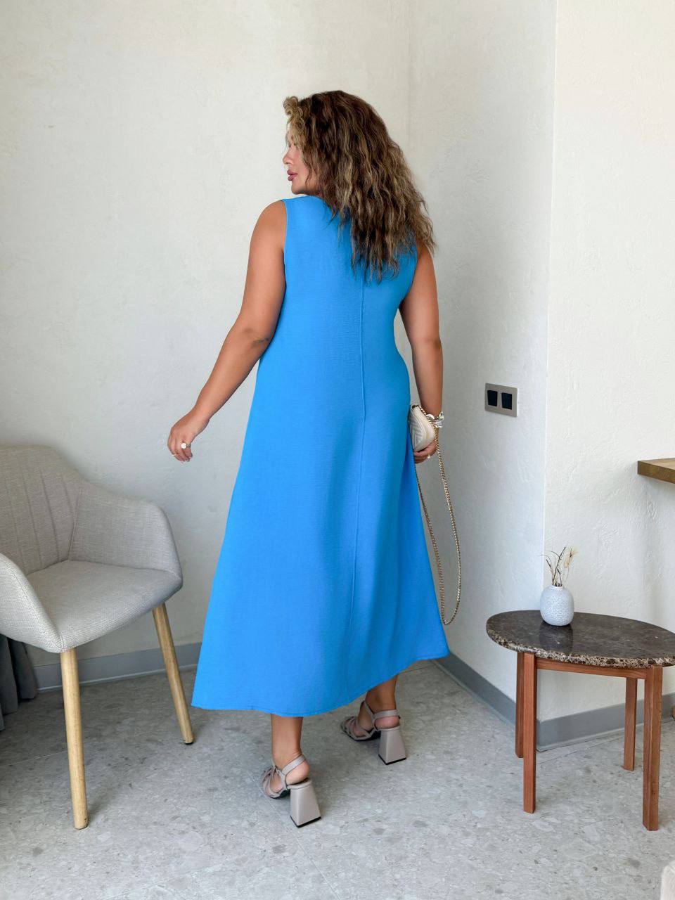 Woman in a blue dress standing in a room with a chair and small table.