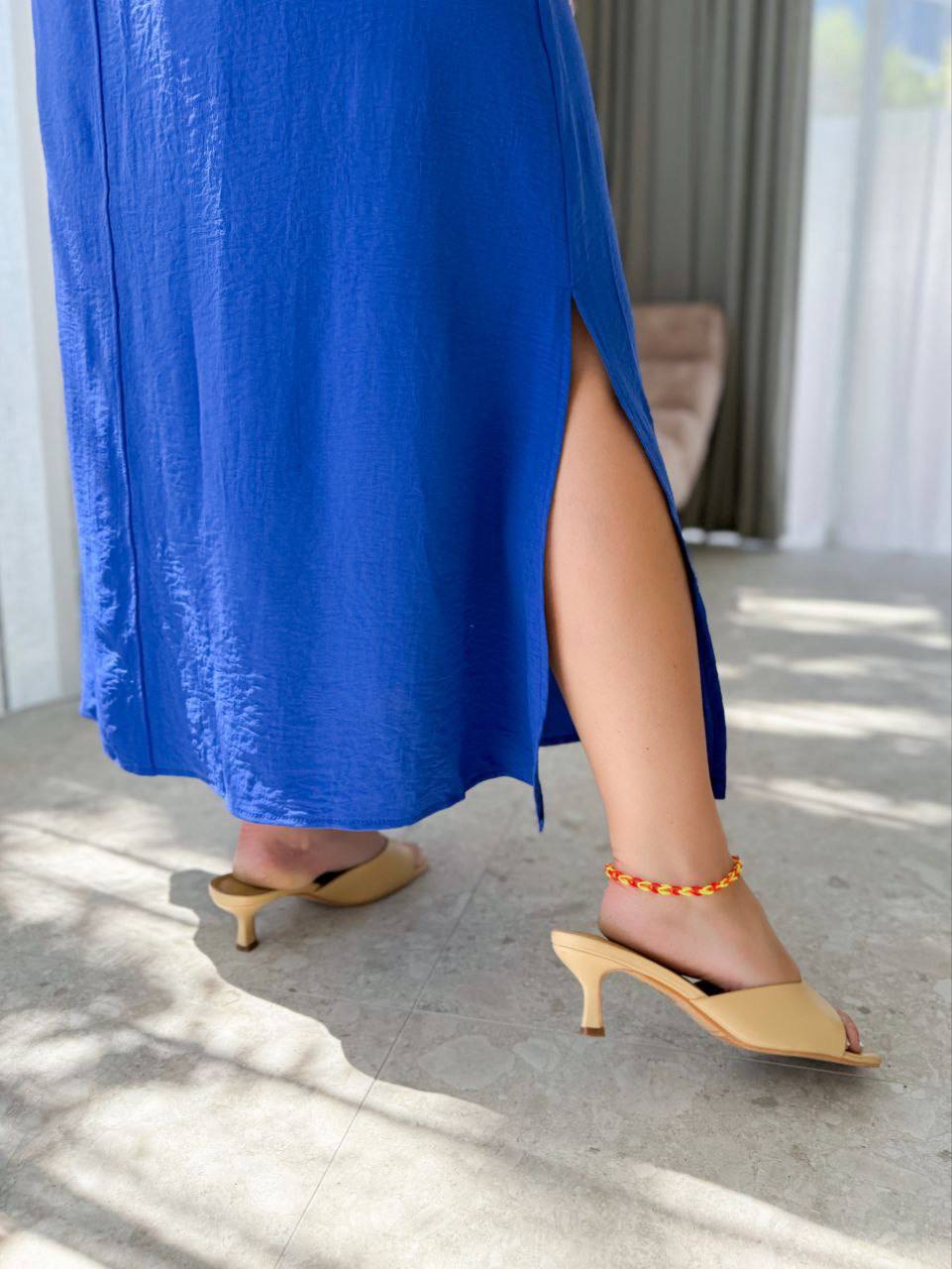 Person wearing a blue dress with a high slit, beige high-heeled shoes, and an orange bracelet on a light-colored floor.