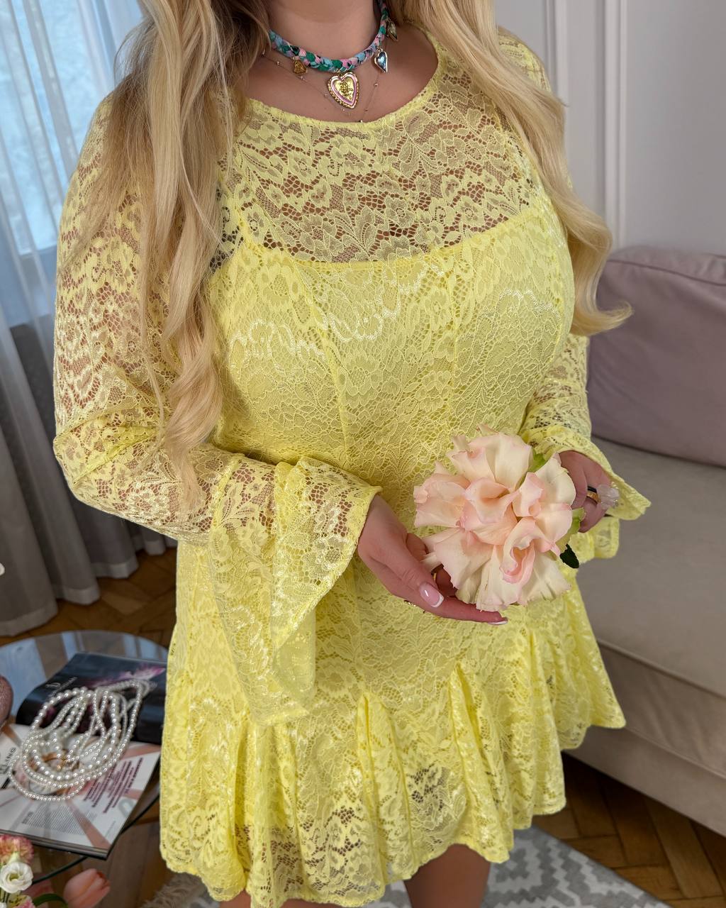 Person wearing a yellow lace dress holding a flower, with a neutral background.