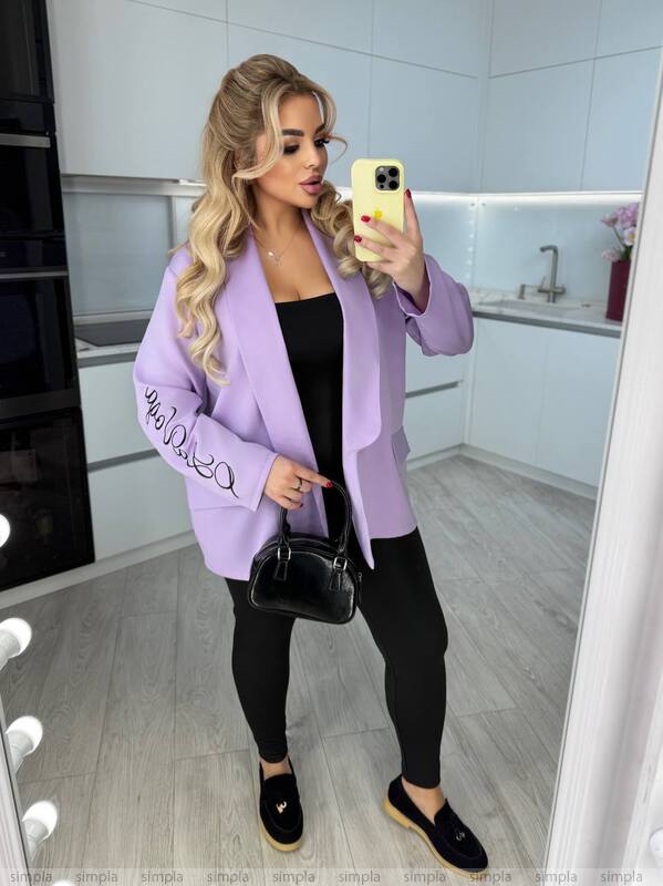 Woman taking a mirror selfie in a kitchen wearing a purple jacket with text, black pants, and black shoes.