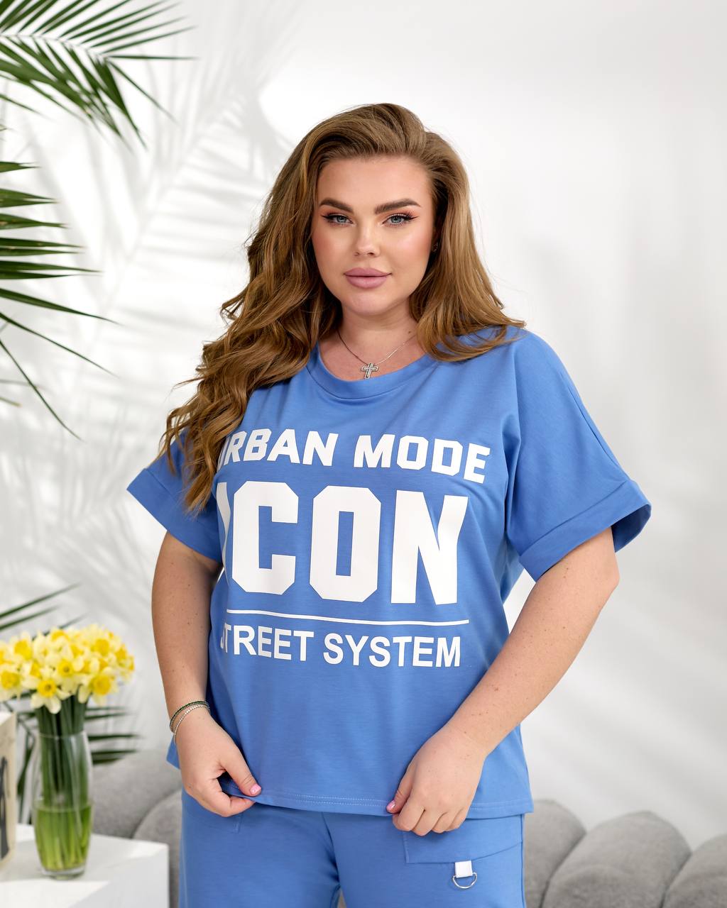 Woman wearing a blue 'Urban Mode Icon Street System' t-shirt in a bright room with plants.