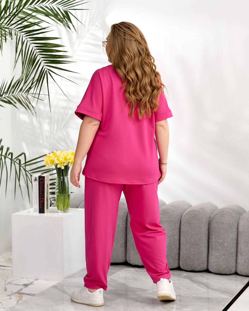 Woman in a pink outfit standing in a modern living room with plants and a gray sofa.