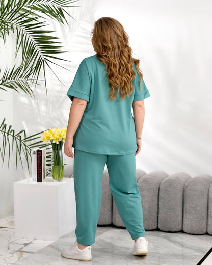 Person wearing a teal outfit standing in a living room with plants and a couch.