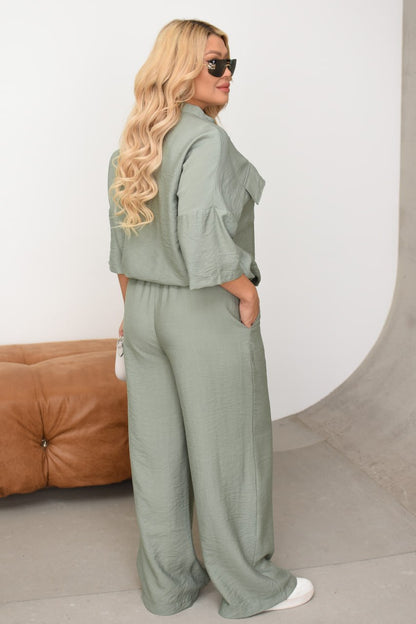 Woman wearing a green outfit standing against a white wall with a brown couch.