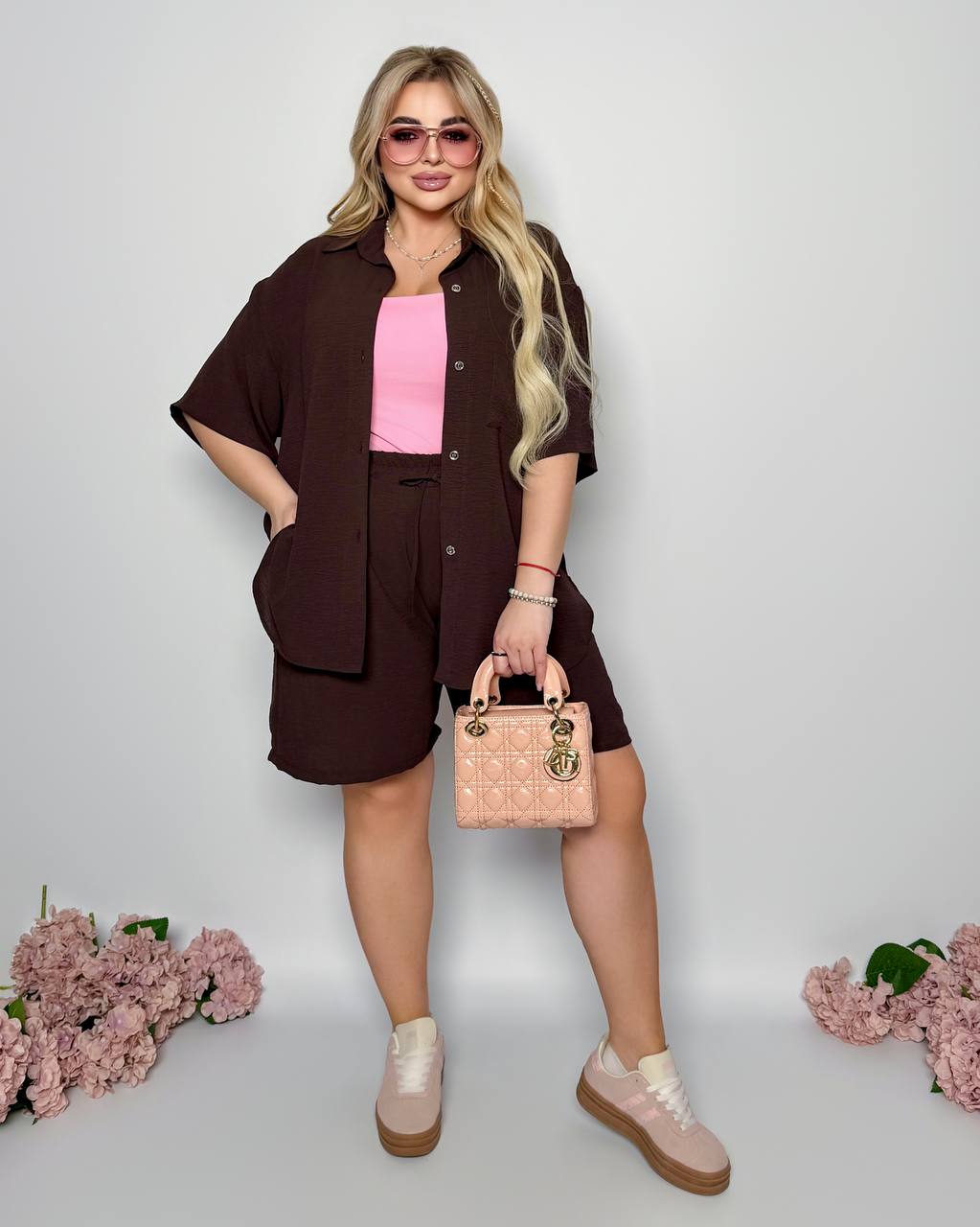 Woman in a brown outfit with pink top and shoes, holding a beige handbag on a light gray background.