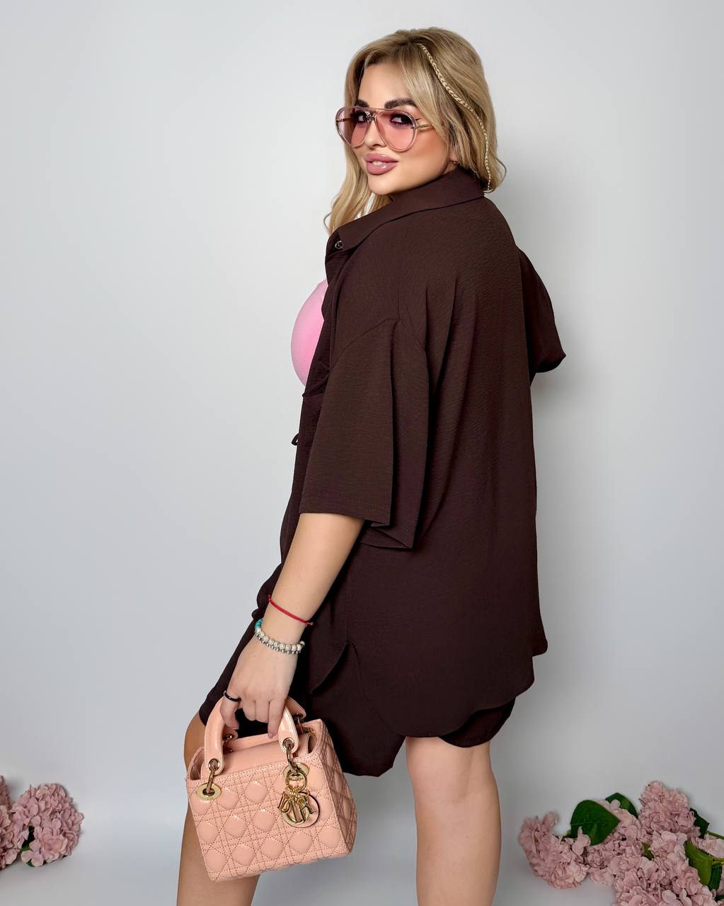 Woman in a brown dress holding a pink handbag against a light gray background with pink flowers.