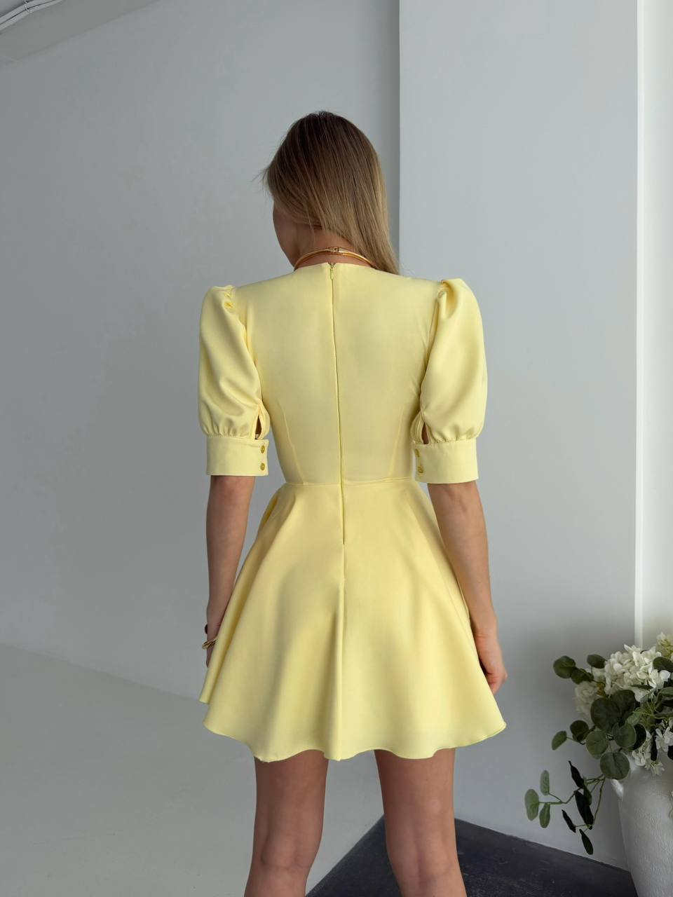 Woman wearing a yellow dress standing against a plain wall with a plant in the corner.
