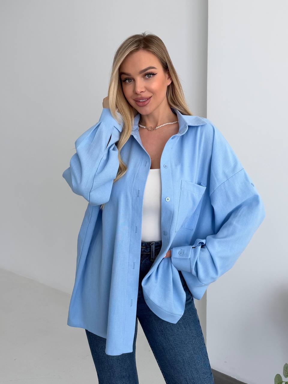 Woman wearing a light blue oversized shirt with a white top and jeans against a plain background