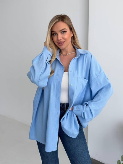 Woman wearing a light blue oversized shirt with a white top and jeans against a plain background