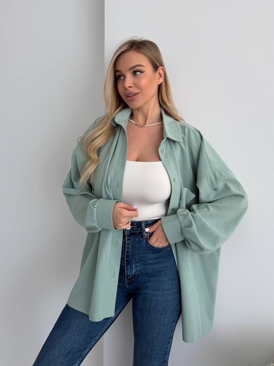 Woman wearing a light green shirt over a white top and blue jeans against a plain background