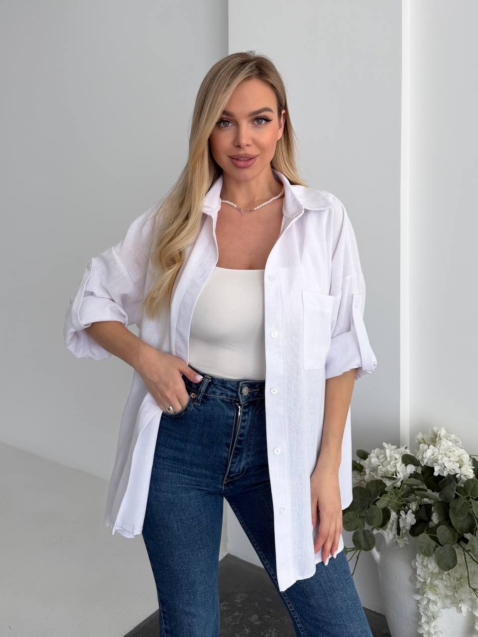 Woman wearing a white shirt and blue jeans standing indoors with a neutral background