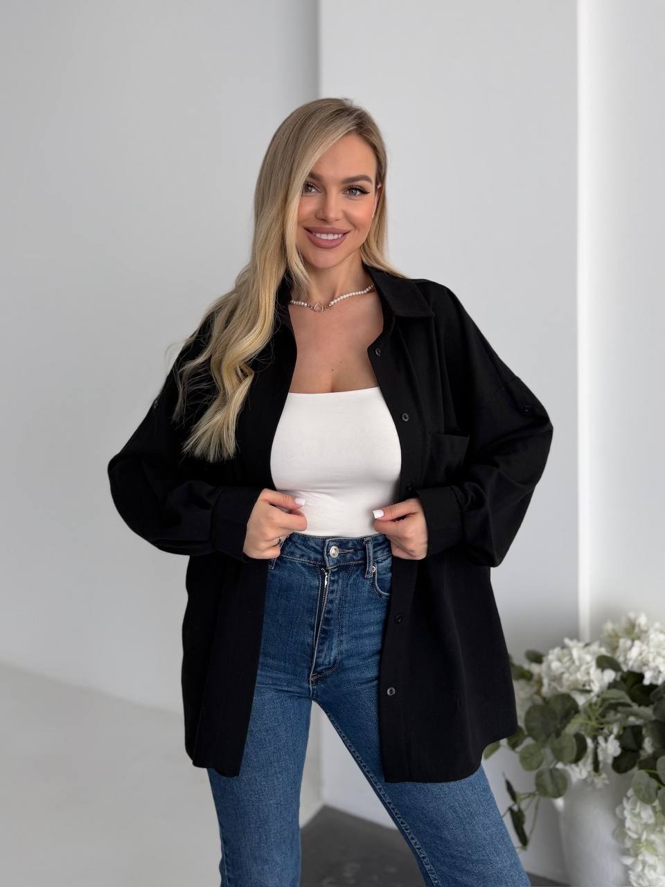 Woman wearing a black jacket, white top, and blue jeans standing indoors with a neutral background.