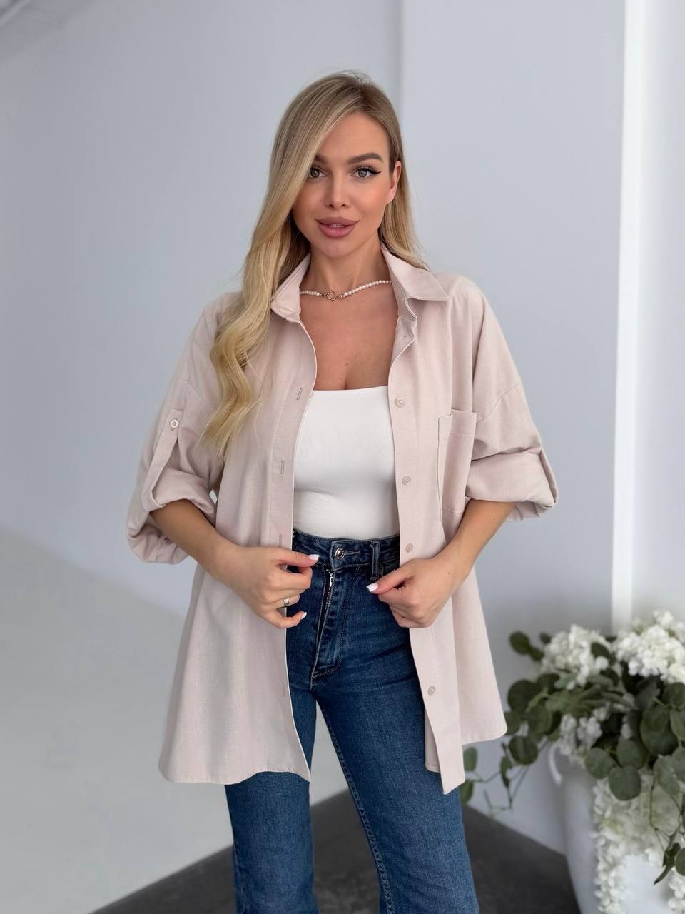 Woman wearing a beige shirt over a white top and blue jeans, standing against a plain background.