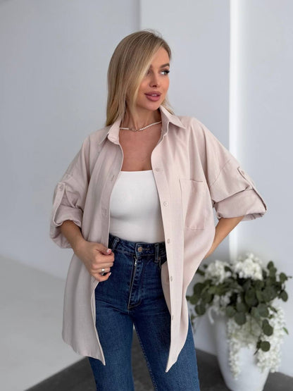 Woman wearing a light pink shirt over a white top and blue jeans, standing indoors with a neutral background.