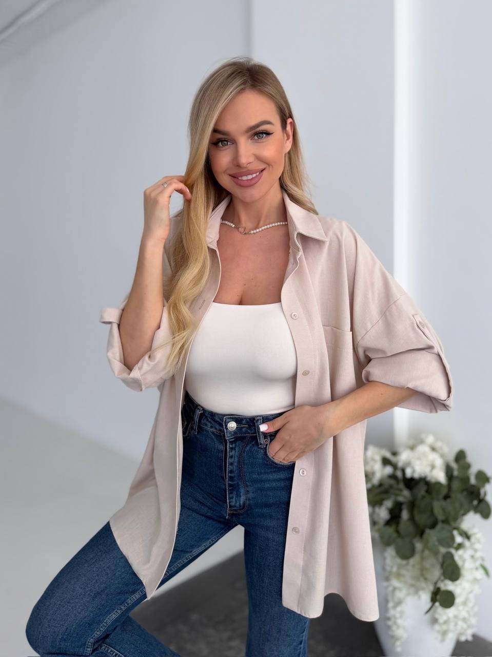 Woman wearing a light pink shirt over a white top and blue jeans, standing indoors with a neutral background.