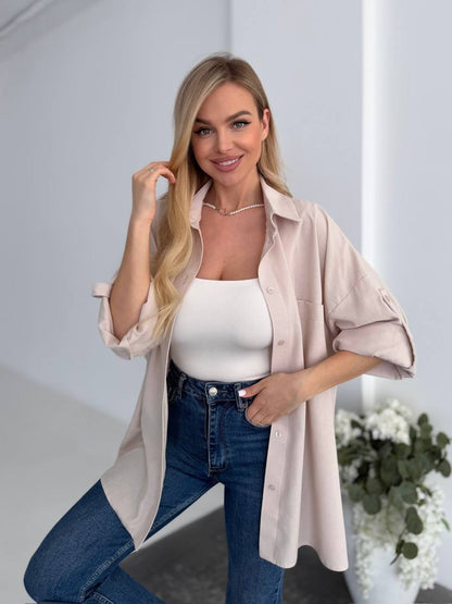 Woman wearing a light pink shirt over a white top and blue jeans, standing indoors with a neutral background.