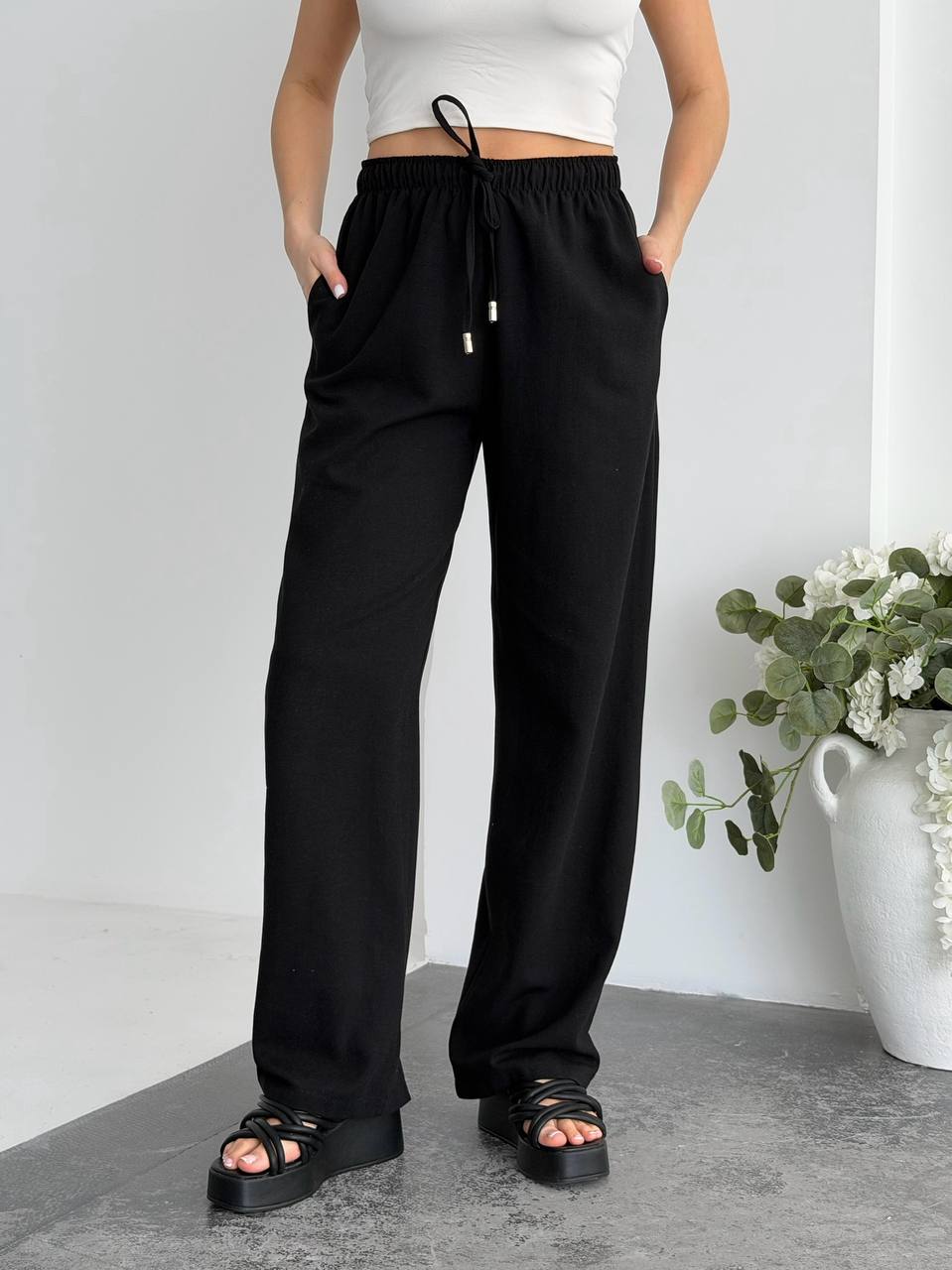 Person wearing black pants with a white top and sandals, standing against a plain background with a vase of flowers.