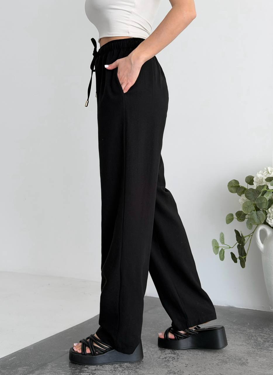 Person wearing black wide-leg pants with a white top and sandals on a light gray background.