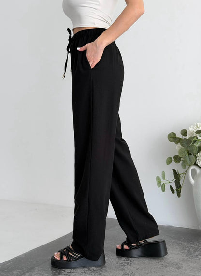 Person wearing black wide-leg pants with a white top and sandals on a light gray background.