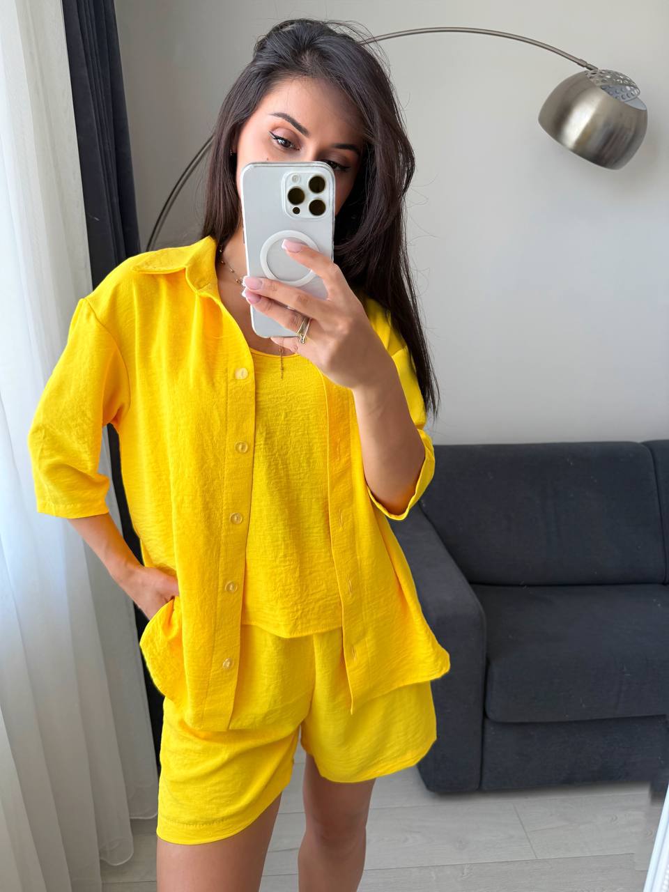 Woman taking a mirror selfie wearing a yellow outfit in a room with a gray sofa and lamp.