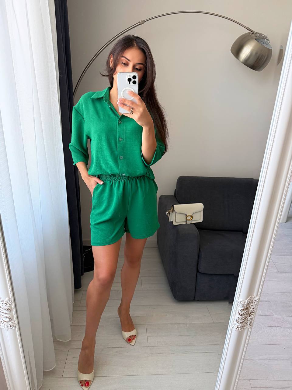Woman taking a mirror selfie wearing a green outfit in a room with a gray sofa and white curtains.