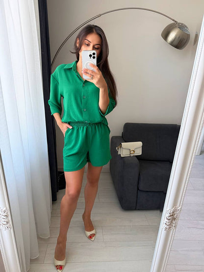 Woman taking a mirror selfie wearing a green outfit in a room with a gray sofa and white curtains.