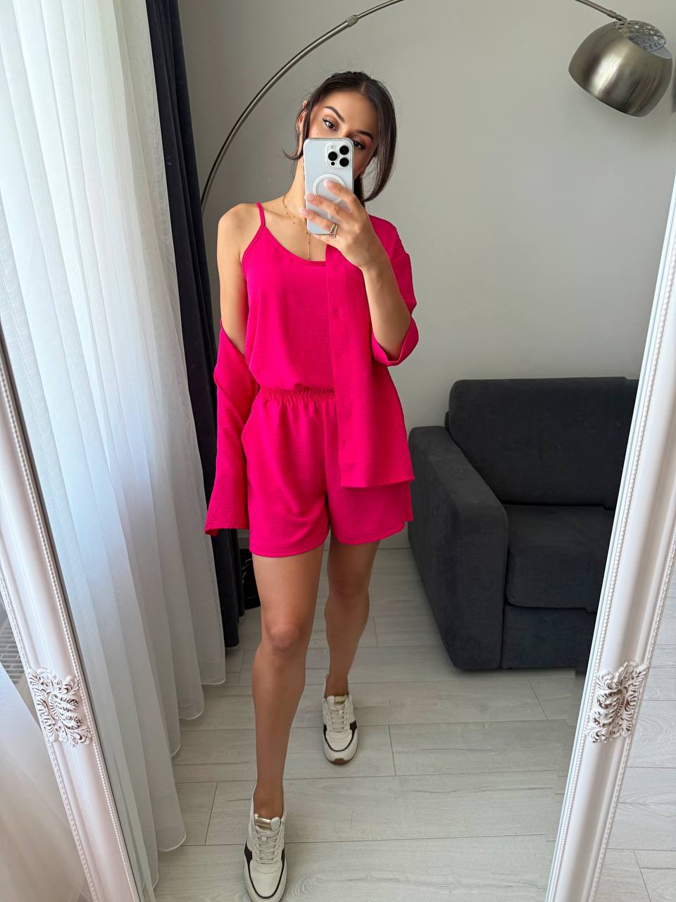 Person wearing a bright pink outfit taking a mirror selfie in a room with a gray sofa.