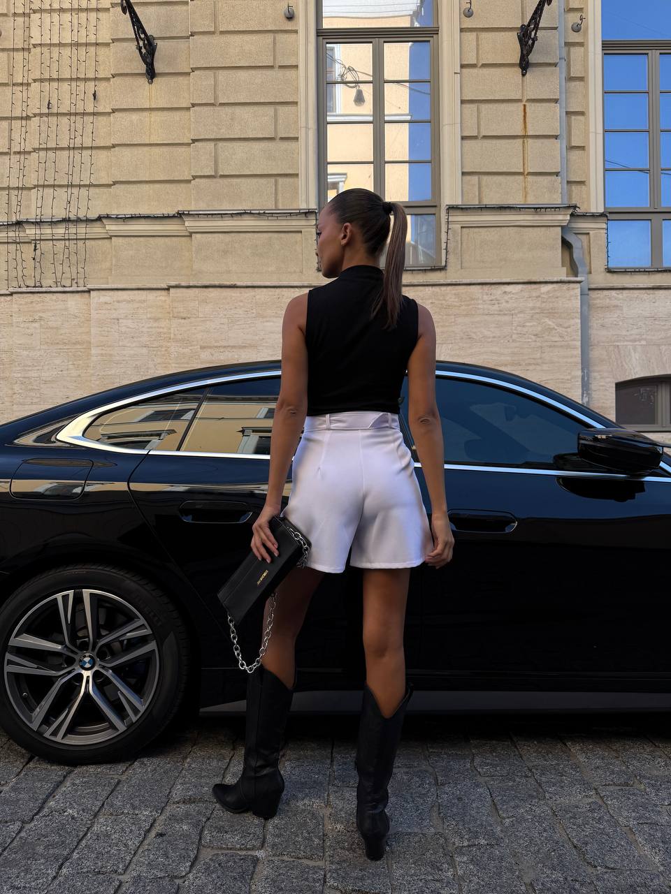 Woman in black sleeveless top and white shorts standing next to a black car on a city street.