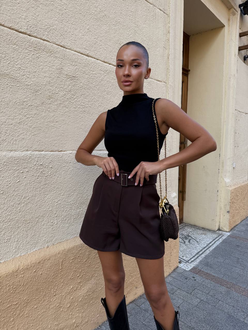 Woman in black sleeveless top and brown shorts standing against a beige wall.