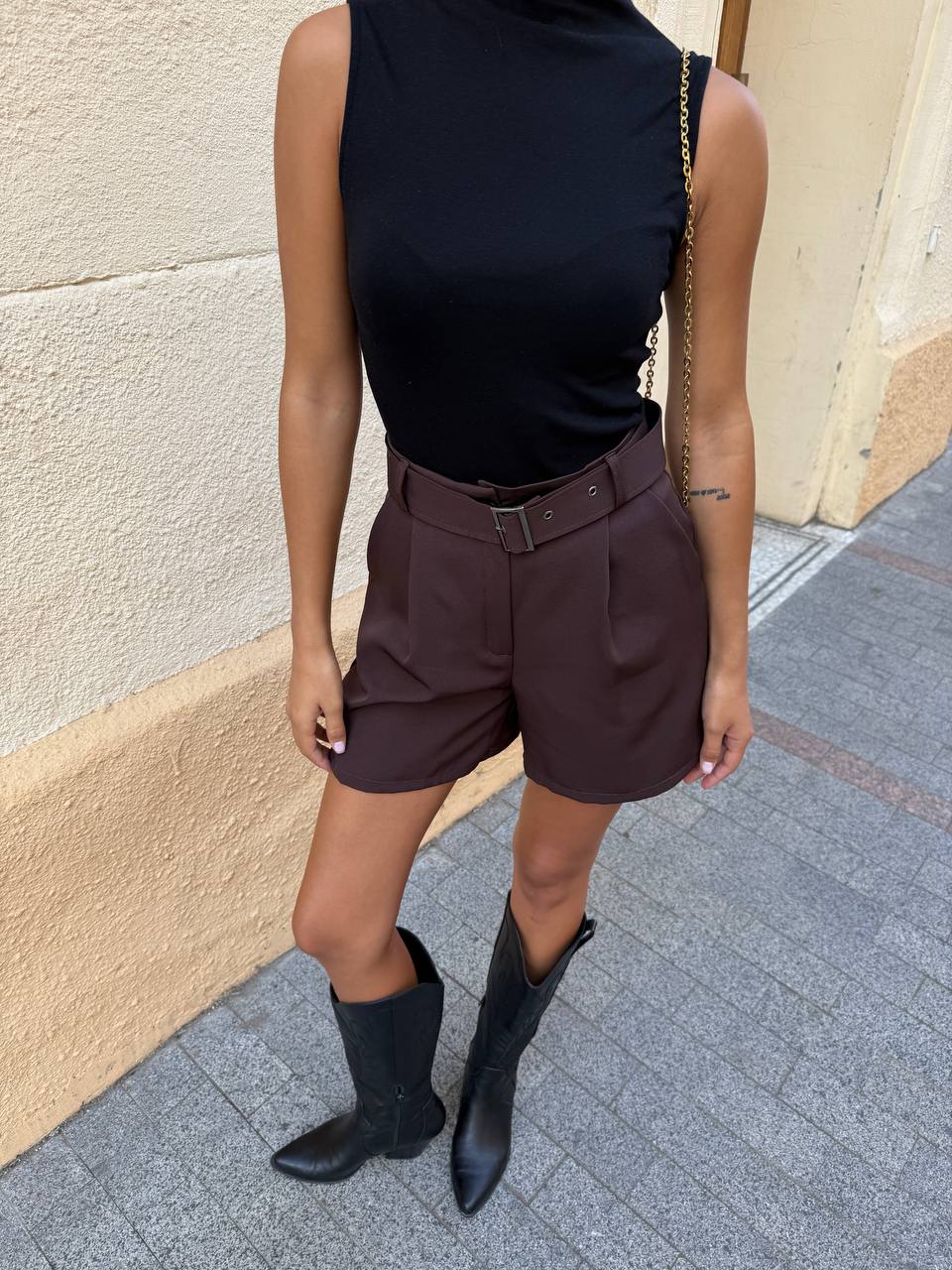 Person wearing a black sleeveless top, brown shorts, and black boots on a sidewalk.