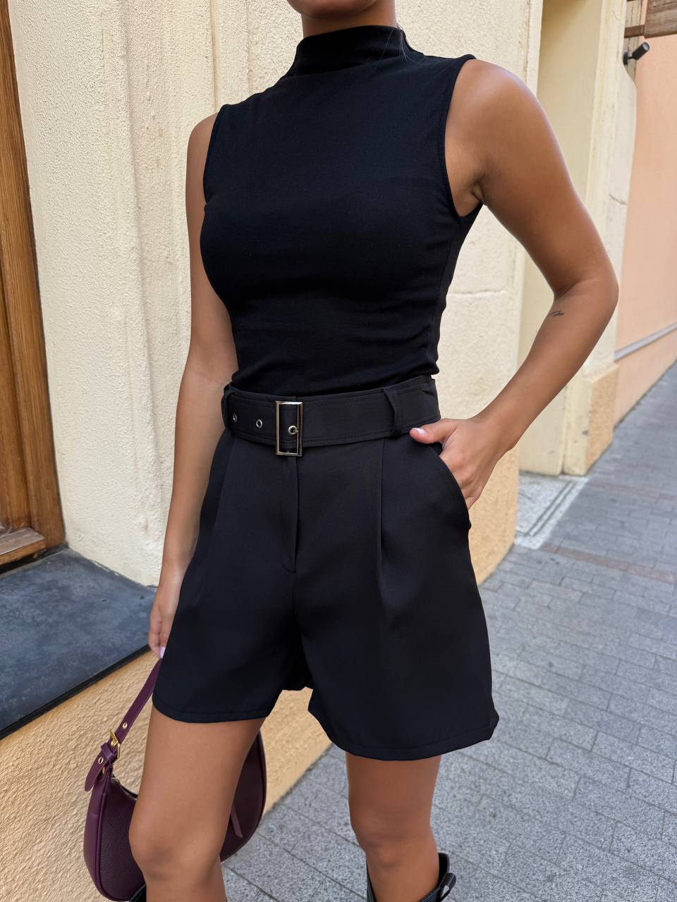 Person wearing a black sleeveless top and shorts with a belt, standing against a beige wall.