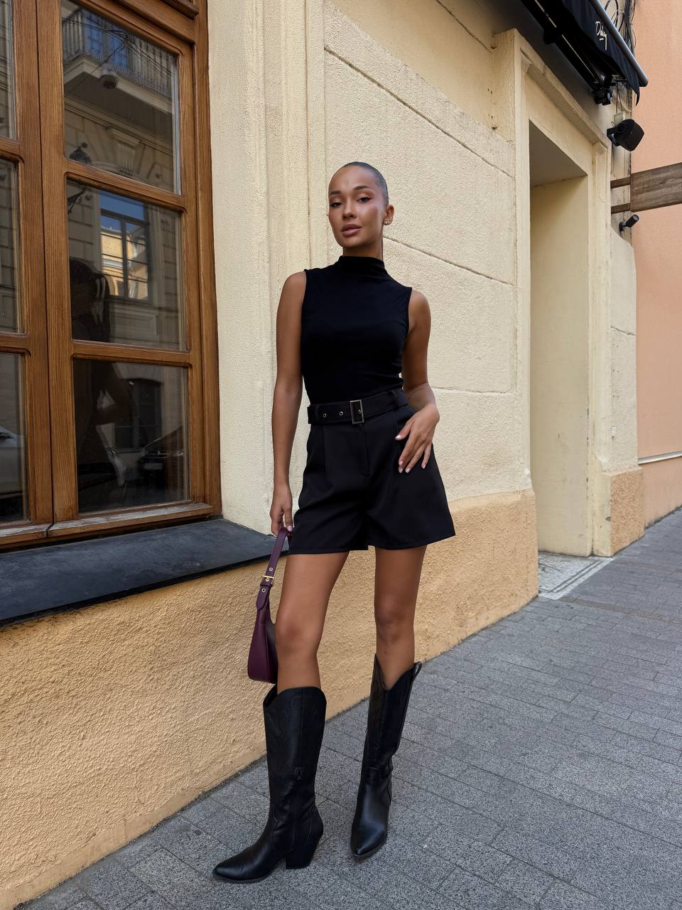Woman in a black sleeveless dress and knee-high boots standing on a street.
