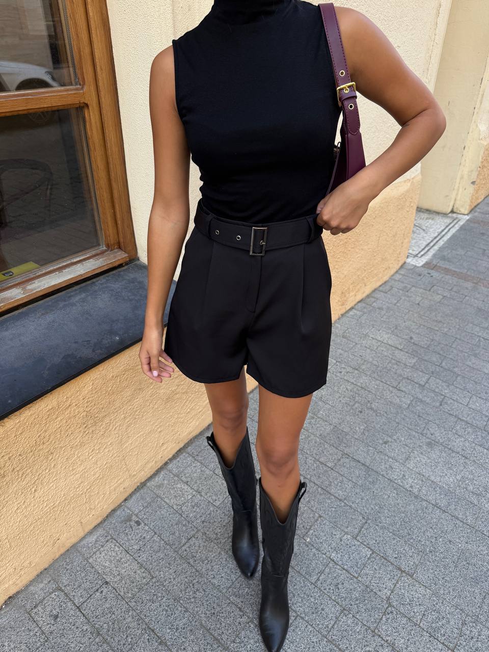 Person wearing a black sleeveless top and shorts with a belt, standing on a sidewalk.