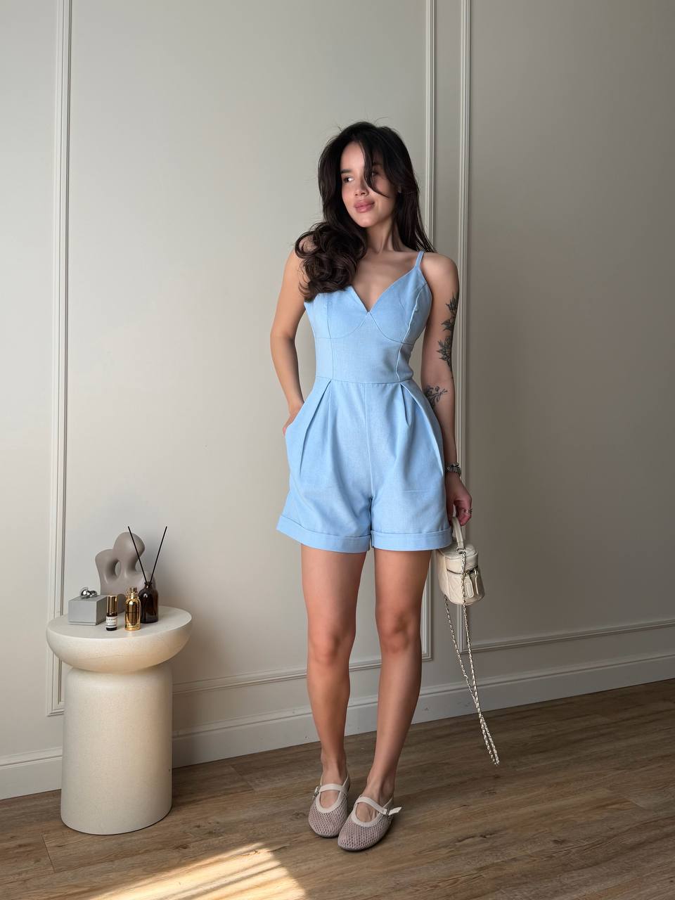 Woman in a light blue romper standing in a room with a white wall and wooden floor.