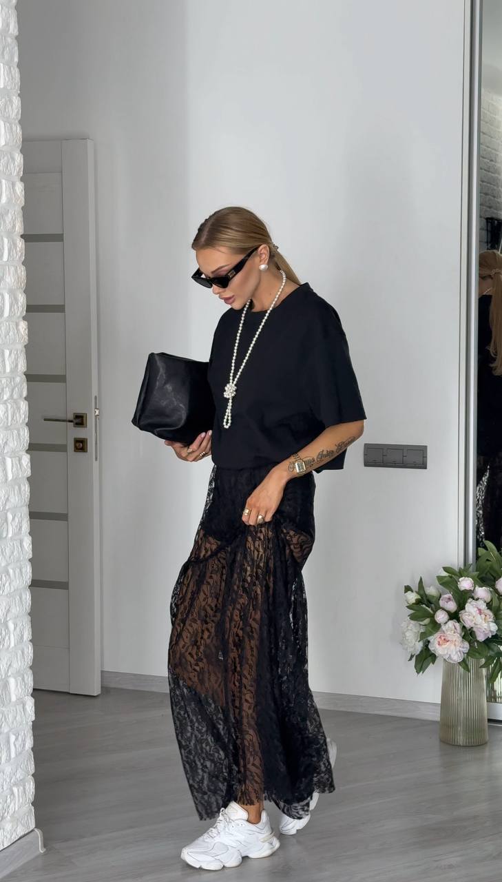 Woman in stylish outfit with black top and lace pants, holding a black clutch.