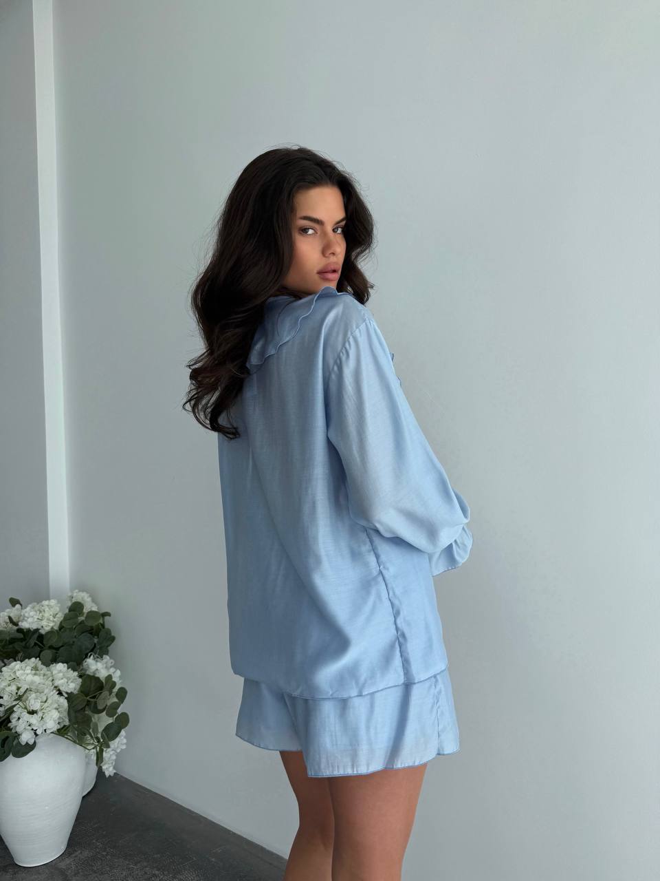 Woman wearing a light blue outfit standing against a plain wall with a vase of flowers on the left.