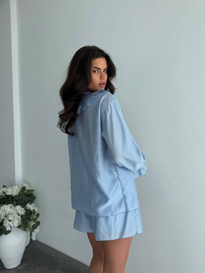 Woman wearing a light blue outfit standing against a plain wall with a vase of flowers on the left.