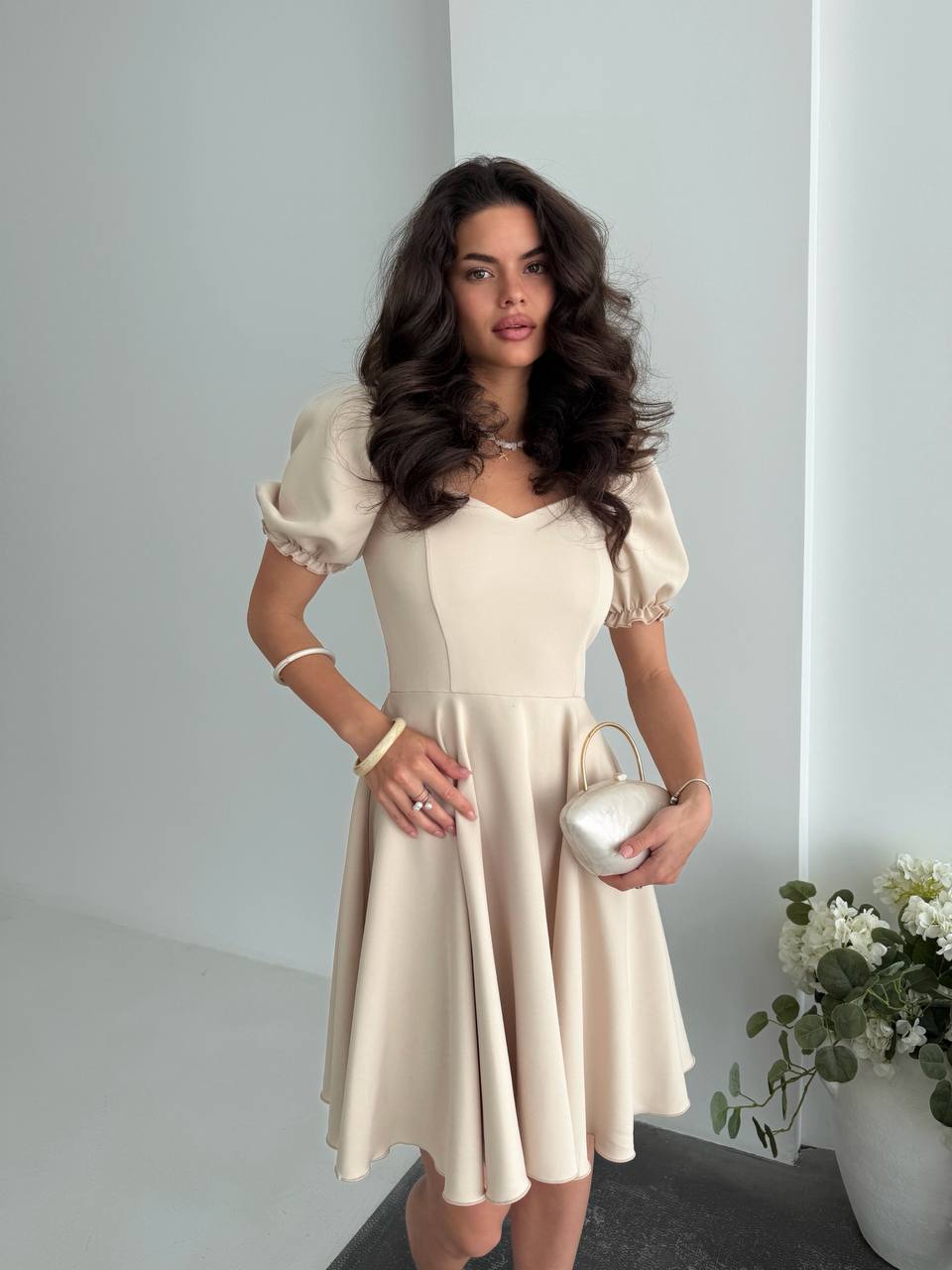 Woman in a beige dress holding a white handbag against a plain background
